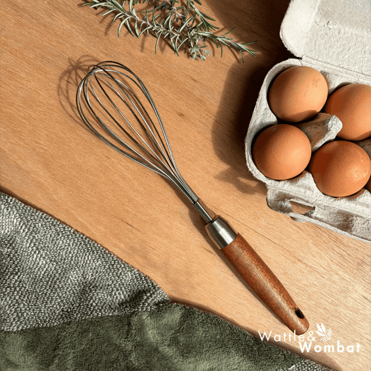 Whisk with wooden handle on a wooden surface next to eggs and rosemary.