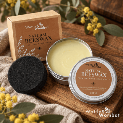Wattle & Wombat Natural Beeswax Premium Wood Care set, featuring an open silver tin with pale yellow beeswax, a black beeswax sponge, and its brown kraft paper box, styled on a wooden tray with linen and yellow flowers.