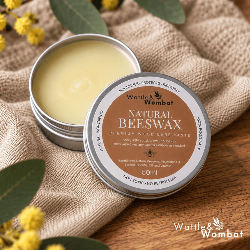 An open round metal tin of Wattle & Wombat Natural Beeswax premium wood care paste sits beside the closed lid on a burlap fabric surface with green leaves and small yellow flowers around it.